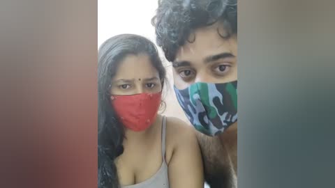 Media: Video of a young woman with long black hair and a red face mask, standing beside a man with curly hair and a green camouflage mask, both smiling.