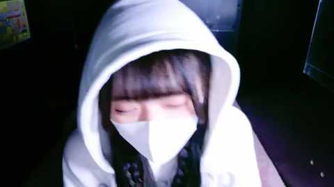 Media: Video of an Asian woman with long black hair, wearing a white hoodie, mask, and sunglasses, sitting in a dimly lit room, eyes closed, looking contemplative.