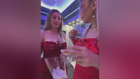 Media: Video of two women, one in a red dress, the other in a red off-shoulder top, toasting with champagne glasses in a dimly lit, modern room with blue lighting and chandeliers.