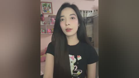 Media: Video of an Asian woman with long black hair, fair skin, and a slender figure, wearing a black T-shirt with a cartoon cat graphic, standing in a pink-themed bedroom with shelves and toys in the background.