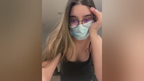 Media: Video of a young Caucasian woman with long, straight brown hair, wearing glasses, a blue mask, and a black tank top. She's leaning forward, hand on her forehead, in a dimly lit bathroom.