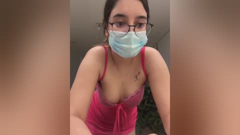 Media: A video shows a young woman with light brown skin, wearing a pink lace camisole, blue mask, and glasses, leaning forward. Her chest is partially visible. Background is blurred.