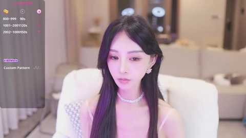 Media: Video of an East Asian woman with long black hair, wearing a white dress, sitting in a modern living room with a beige couch and glass coffee table.