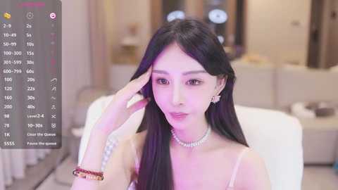 Media: Video of a young East Asian woman with long black hair, wearing a white spaghetti-strap top, pearl necklace, and bracelets. She's in a modern living room with beige walls and a sofa.