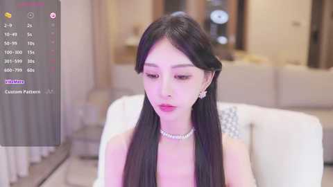 Media: Video of an East Asian woman with long black hair and fair skin, wearing a pearl necklace and a white dress, sitting in a modern, softly lit living room.