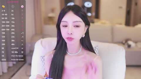 Media: A video of an East Asian woman with long black hair and fair skin, wearing a white off-shoulder dress, sitting in a modern living room with soft lighting.