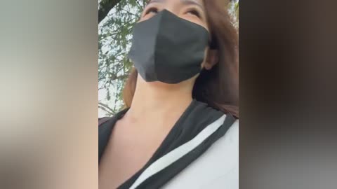 Media: A close-up video of a person wearing a black face mask, a black and white striped shirt, and a shoulder bag, with blurred green foliage in the background.