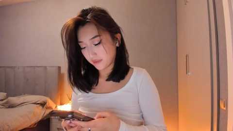 Media: Video of an Asian woman with shoulder-length black hair, wearing a white long-sleeve top, looking down at a smartphone, sitting in a modern, dimly-lit bedroom with beige walls and a gray upholstered headboard.