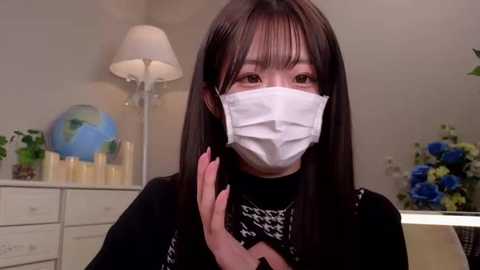 Media: Video of an East Asian woman with long black hair, wearing a white face mask, black top, and pink nails, in a modern indoor setting with a white dresser, globe, and blue flowers.