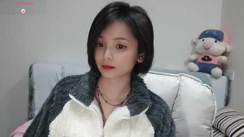 Media: Video of an East Asian woman with short black hair, fair skin, and red lipstick, wearing a gray and white fleece jacket, sitting on a white couch with a plush toy in the background.