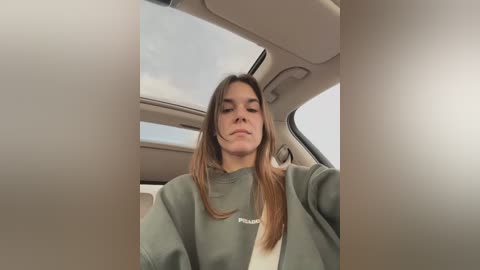 Media: Video of a young woman with long, straight brown hair, wearing a green sweatshirt, seated in a car with beige interior, smiling slightly.