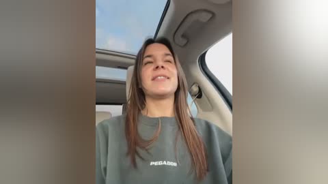 Media: Video of a young woman with long brown hair, wearing a grey \"PEACE\" sweatshirt, smiling, sitting in a car with a sky-blue sky visible through the sunroof.