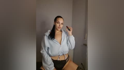 Media: Video of a light-skinned woman with a medium build, long dark hair, wearing a light blue hoodie and black lace underwear, holding a phone, in a dimly lit room.