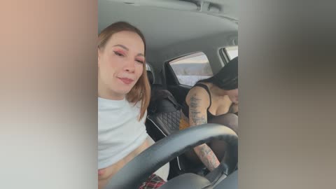 A video of a young woman with fair skin, light brown hair, and pink lipstick, wearing a white crop top and black bra, driving a car with a tattooed passenger in the backseat.