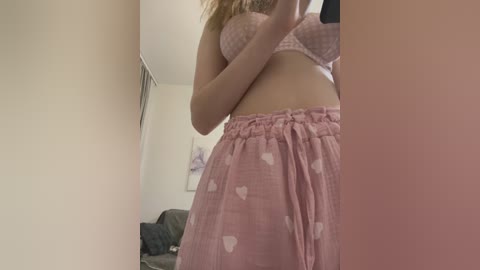 Media: Video of a fair-skinned woman with blonde hair in pink lingerie, a bra and heart-patterned skirt, capturing a mirror reflection in a dimly lit bedroom.