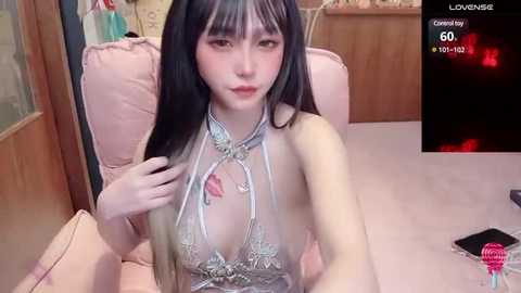 Media: A young Asian woman with long black hair and fair skin, wearing a revealing silver lace lingerie set, sits on a plush pink chair in a cozy room, with a smartphone displaying a \"Camgirl\" label and stats in the background.