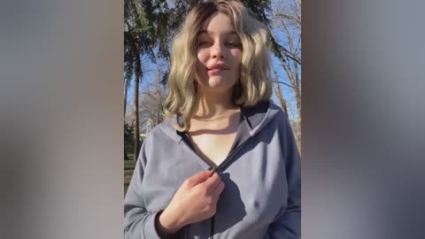 Media: Video of a young, fair-skinned woman with shoulder-length blonde hair, wearing a gray hoodie, standing outdoors with a clear blue sky and trees in the background.