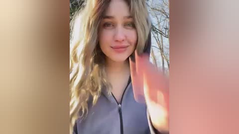 Media: Video of a fair-skinned woman with wavy blonde hair, smiling, wearing a grey zip-up jacket, partially obscured by a person's hand on the right. Background includes blurred trees.