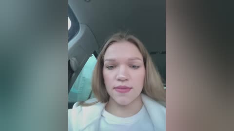 Media: Video of a young Caucasian woman with fair skin, straight blonde hair, wearing a white sweater, sitting in a car.