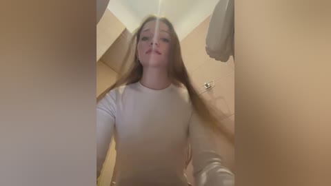 Media: Video of a young woman with long, straight blonde hair, wearing a white long-sleeved top, standing in a narrow hallway with beige walls.