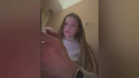 Media: Video of a young woman with long, straight brown hair, wearing a white top, standing in a narrow hallway with beige walls, looking up with a slightly worried expression.