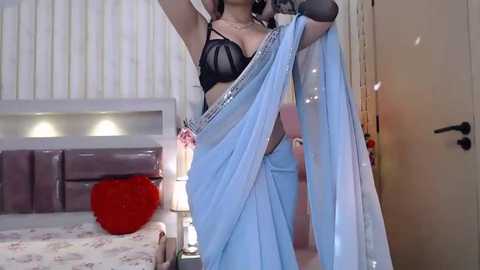 Media: A video of a woman in black lingerie and a light blue saree, posing in a bedroom with a bed, red heart pillow, and white walls.