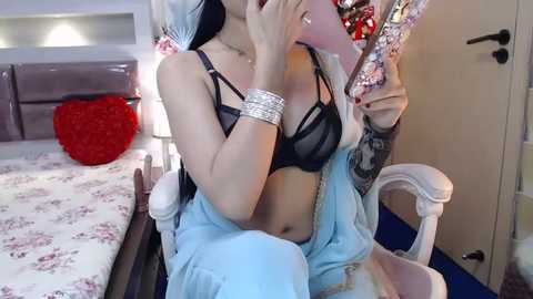 Media: A video of a fair-skinned woman with long, platinum blonde hair, wearing a black lace bra, light blue pants, and a pink phone case. She sits on a white chair with intricate floral wallpaper and a red heart pillow in the background.