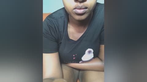 Media: Video of a young Black woman with medium skin tone, wearing a black T-shirt with a small, white cartoon dog, sitting with arms crossed, mouth slightly open, in a dimly lit room.