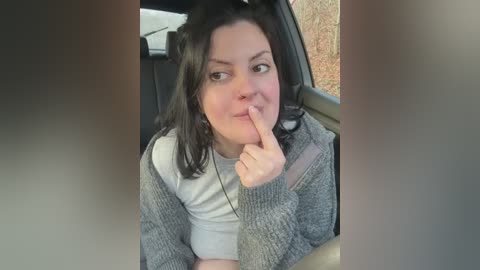 Media: Video of a young woman with shoulder-length black hair, light skin, and freckles, wearing a gray knitted cardigan over a white shirt, sitting in a car, with a finger to her lips in a shushing gesture.