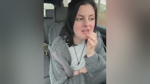 Media: A video of a young woman with fair skin and shoulder-length dark hair, wearing a gray cardigan and a white t-shirt, sitting in a car with a neutral expression and hand near her mouth.