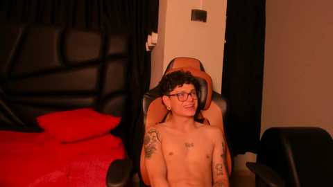 A video of a shirtless, tattooed, slender, young man with glasses sitting in a black gaming chair, surrounded by a dimly lit bedroom with a red bedspread and dark curtains.