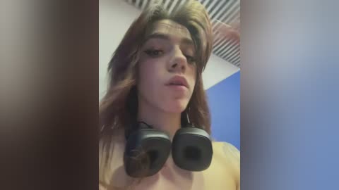 Media: Video of a young person with long, wavy brown hair and a fair complexion, wearing black headphones, in a dimly lit room with blue and white walls.