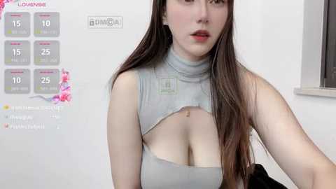 Media: A video of a fair-skinned woman with long brown hair, wearing a revealing light grey crop top, exposing ample cleavage, in a modern, minimalistic room with a calendar displaying dates and events.