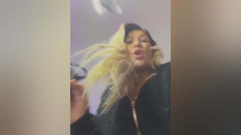 Media: Video of a young, light-skinned woman with long, voluminous blonde hair and a black jacket, capturing a moment of joy and movement.