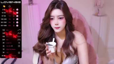 Media: A video of a young Asian woman with long, wavy brown hair, wearing a strapless silver dress, holding a white cup, in a softly lit, pink-hued room.