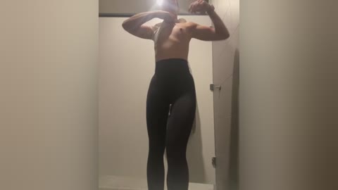 Media: Video of a topless, fit, light-skinned woman with small breasts, wearing black leggings, standing in a changing room, holding a phone to her face, capturing a mirror reflection.