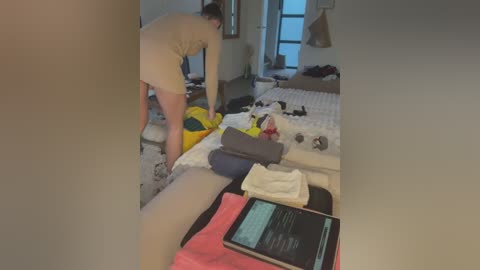 Media: Video of a person in beige underwear and socks, bending over to put on socks, in a messy bedroom with a bed, clothes, and a tablet.