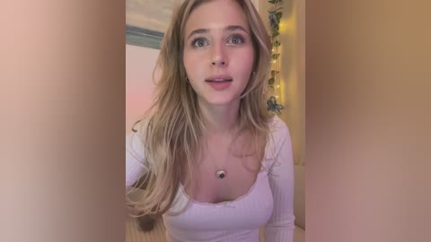 Media: Video of a young, fair-skinned, blonde woman with blue eyes, wearing a white, long-sleeve, low-cut top, standing in a warmly lit room with beige walls and green curtains.