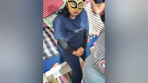 Media: A video of a woman in a blue shirt and yellow mask standing amidst colorful, patterned blankets in a cluttered room.