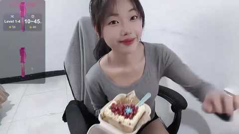 Media: A video of an East Asian woman in a gray top, seated in an office chair, holding a sandwich with blueberries. The background shows a white wall with a clock and a pink marker.