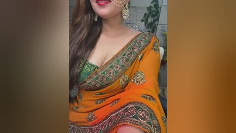 Media: Video of a South Asian woman in an orange sari with intricate golden embroidery, wearing green blouse, gold earrings, and a nose ring. Background features blurred outdoor elements.