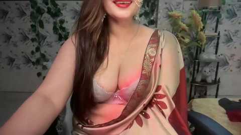 Media: Video of a smiling, fair-skinned woman with long brown hair, wearing a revealing, beige and red sari with a pink lace bra, against a floral wallpaper background.