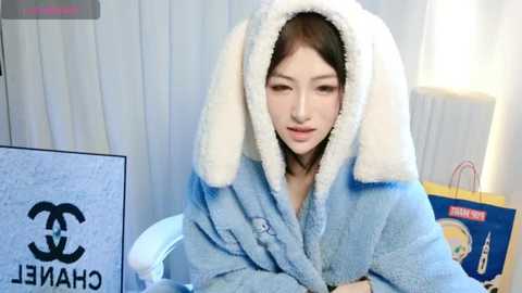 Media: Video of a young Asian woman with straight black hair, wearing a light blue Chanel robe with white faux-fur hood, seated in a bright, modern room with white curtains.