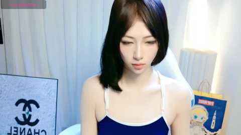 Media: Video of an East Asian woman with straight black hair, wearing a blue tank top with white straps, sitting in a white room with vertical blinds, lamp, and a bag.