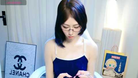 Media: Video of an East Asian woman with shoulder-length black hair, wearing glasses and a dark blue camisole, sitting in a modern room with white walls and a Chanel poster, surrounded by colorful shopping bags.