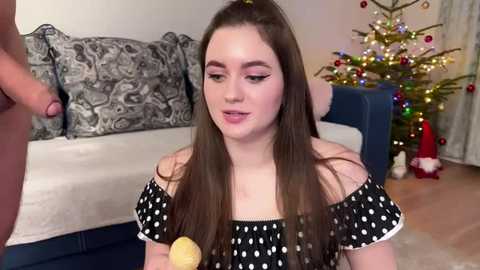 Media: A video of a young woman with long brown hair, fair skin, and dark eyeliner, kneeling in a living room, applying makeup to a man's erect penis. Christmas tree and decorations in background.