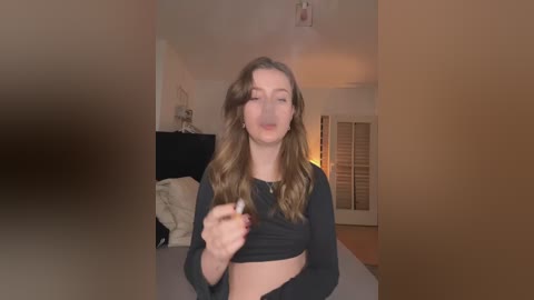 Video of a young woman with long brown hair, wearing a black crop top, standing in a dimly lit bedroom with white walls and a bed with white sheets in the background.