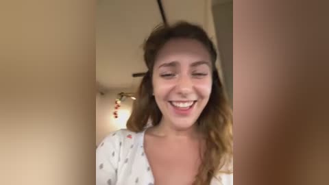 Media: Video of a young woman with light skin and wavy brown hair, smiling, wearing a white top with small print, standing in a dimly lit room with beige walls and a hanging light fixture.