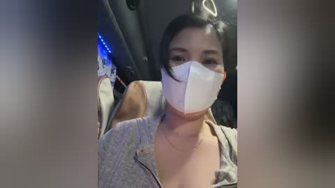 Media: Video of a woman with light brown skin, dark hair, and a light gray jacket, wearing a white face mask, sitting in a car with dim lighting and blue LED lights.