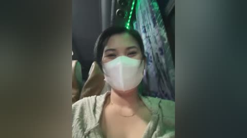 Media: Video of a young Asian woman with straight black hair, wearing a white surgical mask and a light green cardigan, standing indoors with dim lighting and green string lights in the background.